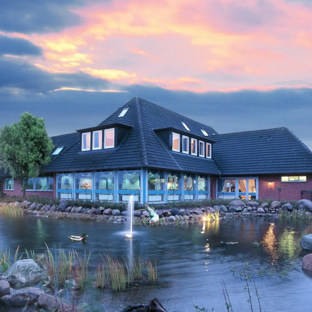 Hochzeitslocation - Das Bild zeigt ein großes, einstöckiges Gebäude mit einem Satteldach aus dunklen Ziegeln. Es wirkt modern und gepflegt. Der Gebäudestill ist im Stil eines Landhauses gehalten, aber mit modernen Elementen. Große Fensterfronten, die sich über einen Teil der Länge des Gebäudes erstrecken, lassen den Innenraum hell und einladend erscheinen. Vor dem Haus befindet sich ein Teich, der von Steinen und Wasserpflanzen umgeben ist. Im Teich ist ein kleiner Springbrunnen zu sehen. Zwei Enten schwimmen auf dem Wasser. Der Himmel ist mit rosa und violetten Farbtönen der Dämmerung oder Morgendämmerung gefärbt, was eine ruhige und friedliche Stimmung erzeugt. Das Gesamtbild strahlt Ruhe, Behaglichkeit und eine gewisse Eleganz aus. Es könnte sich um ein Hotel, ein Restaurant oder ein ähnliches Gebäude handeln, das auf Entspannung und Wohlbefinden ausgerichtet ist. Die Beleuchtung suggeriert Abendstunden oder frühen Morgen. Die Landschaftsgestaltung mit Teich und Enten wirkt harmonisch integriert und unterstreicht den ruhigen Charakter des Ortes. - SOLUNA ELEMENTS