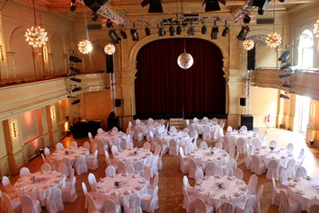 Hochzeit: Festlicher Ballsaal mit runden Tischen und Bankett-Bestuhlung mit weißen Stuhl-Hussen - Heimathafen Neukölln