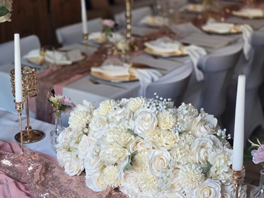 Hochzeitslocation: Blick vom Brauttisch im festlich geschmückten Saal des Wanderheim Germersheim. Ein üppiges Blumengesteck aus weißen und cremefarbenen Rosen ziert den Tisch, ergänzt durch goldene Kerzenhalter und roségoldene Akzente. Die harmonische Kombination aus Farben und Licht verleiht der Hochzeitsdekoration eine luxuriöse, zugleich warme Atmosphäre – ideal für stilvolle Feiern in der Pfalz. - Wanderheim Germersheim