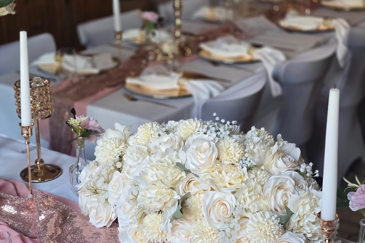 Hochzeitslocation: Blick vom Brauttisch im festlich geschmückten Saal des Wanderheim Germersheim. Ein üppiges Blumengesteck aus weißen und cremefarbenen Rosen ziert den Tisch, ergänzt durch goldene Kerzenhalter und roségoldene Akzente. Die harmonische Kombination aus Farben und Licht verleiht der Hochzeitsdekoration eine luxuriöse, zugleich warme Atmosphäre – ideal für stilvolle Feiern in der Pfalz. - Wanderheim Germersheim
