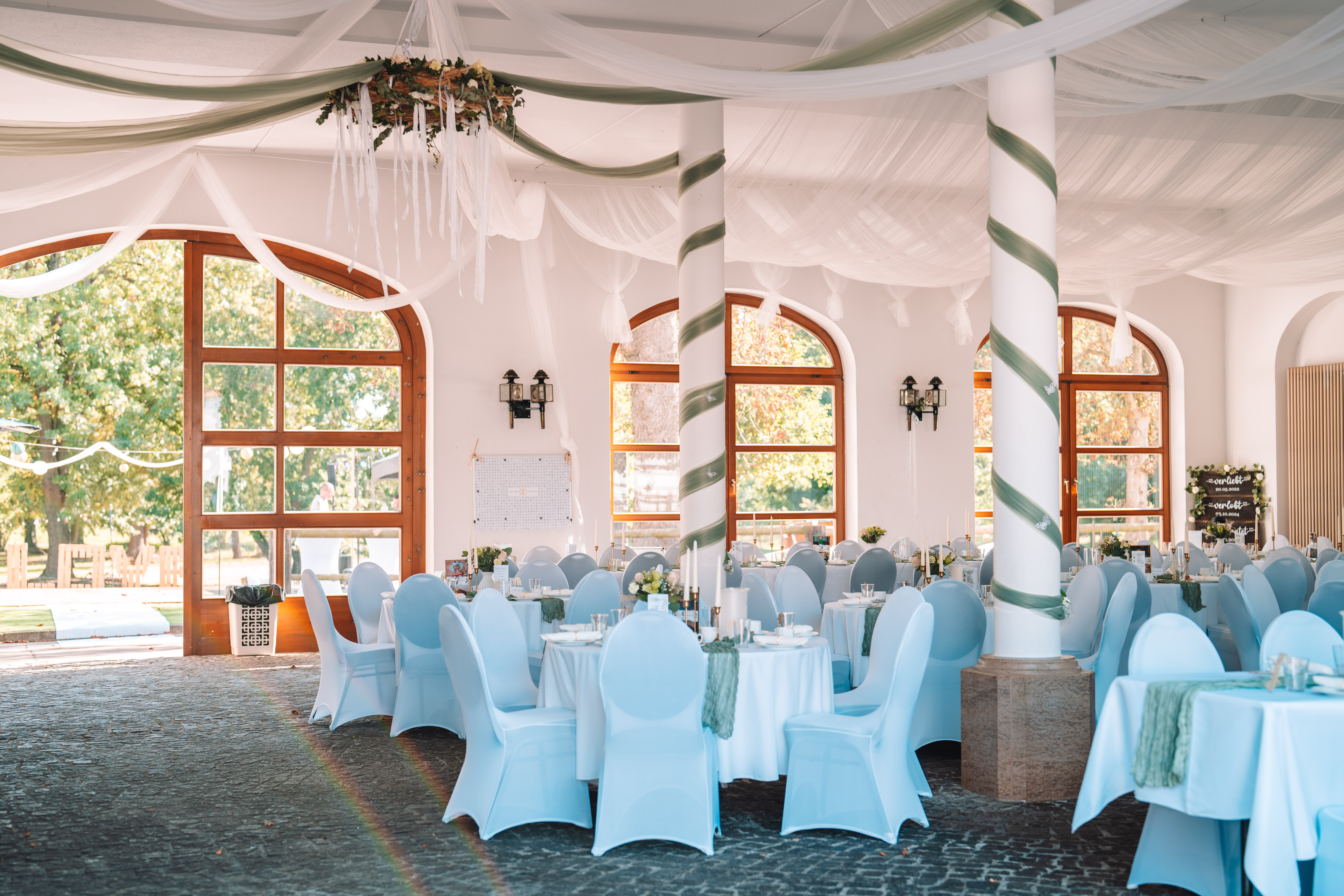Mariage - Bennewitz - Festsaal mit weißen Stuhlhussen und runden, weiß gedeckten Tischen. Deckenverhang aus weißem Stoff mit Blumenkranz, Säulen mit weiß-grünem Band umwickelt. Große Rundbogenfenster mit Blick ins Grüne, Regenbogenreflexion auf dem Steinboden. - Remise am Gestüt Sachsen in Cavertitz