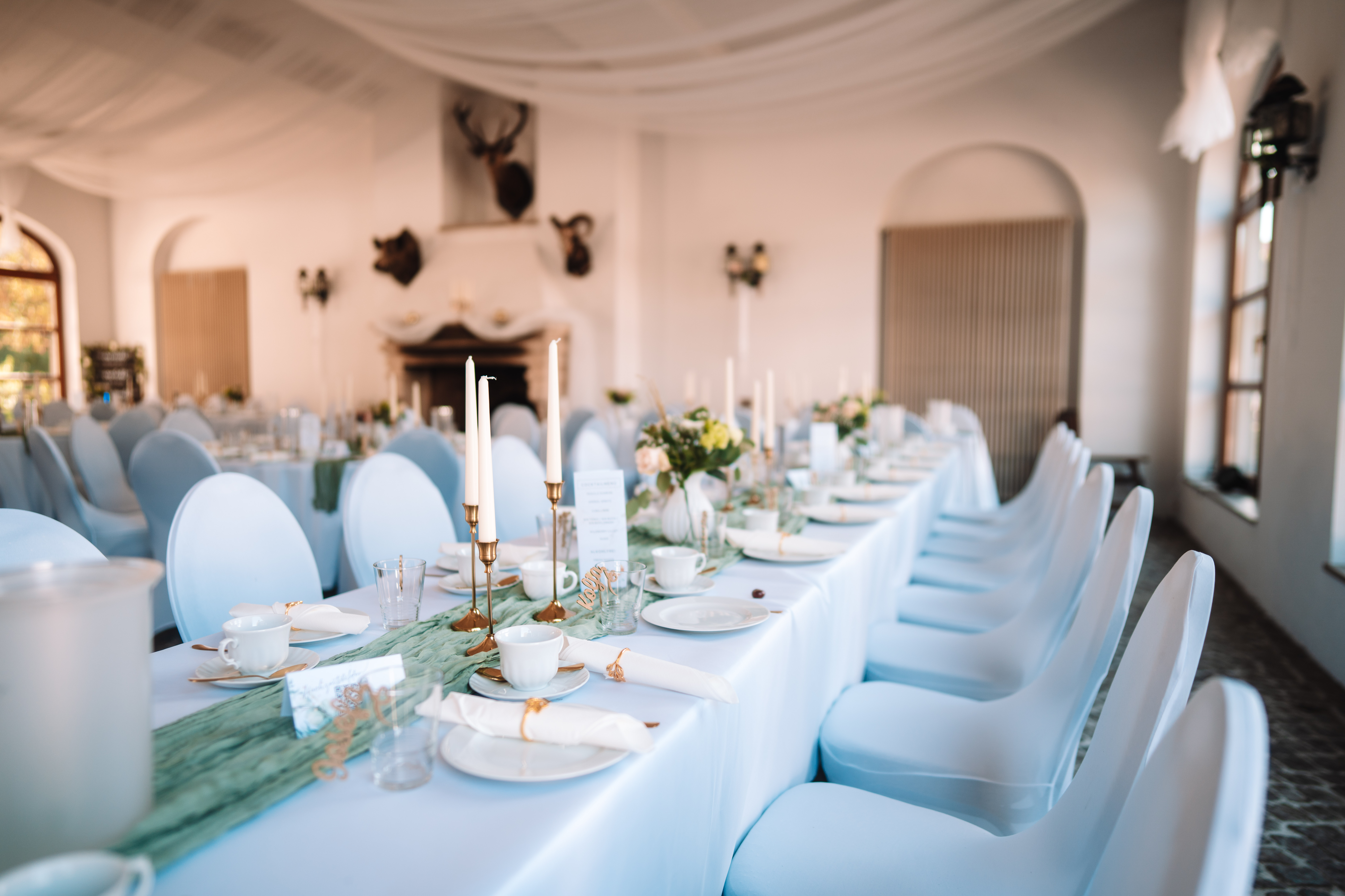 Mariage - Bennewitz - Lange festlich gedeckte Tafel mit weißen Hussenstühlen, goldenen Kerzenleuchtern, weißen Stabkerzen, Blumenvasen und salbeigrünem Tischläufer. Im Hintergrund unscharf ein Kamin mit Jagdtrophäen (Hirschgeweih). - Remise am Gestüt Sachsen in Cavertitz