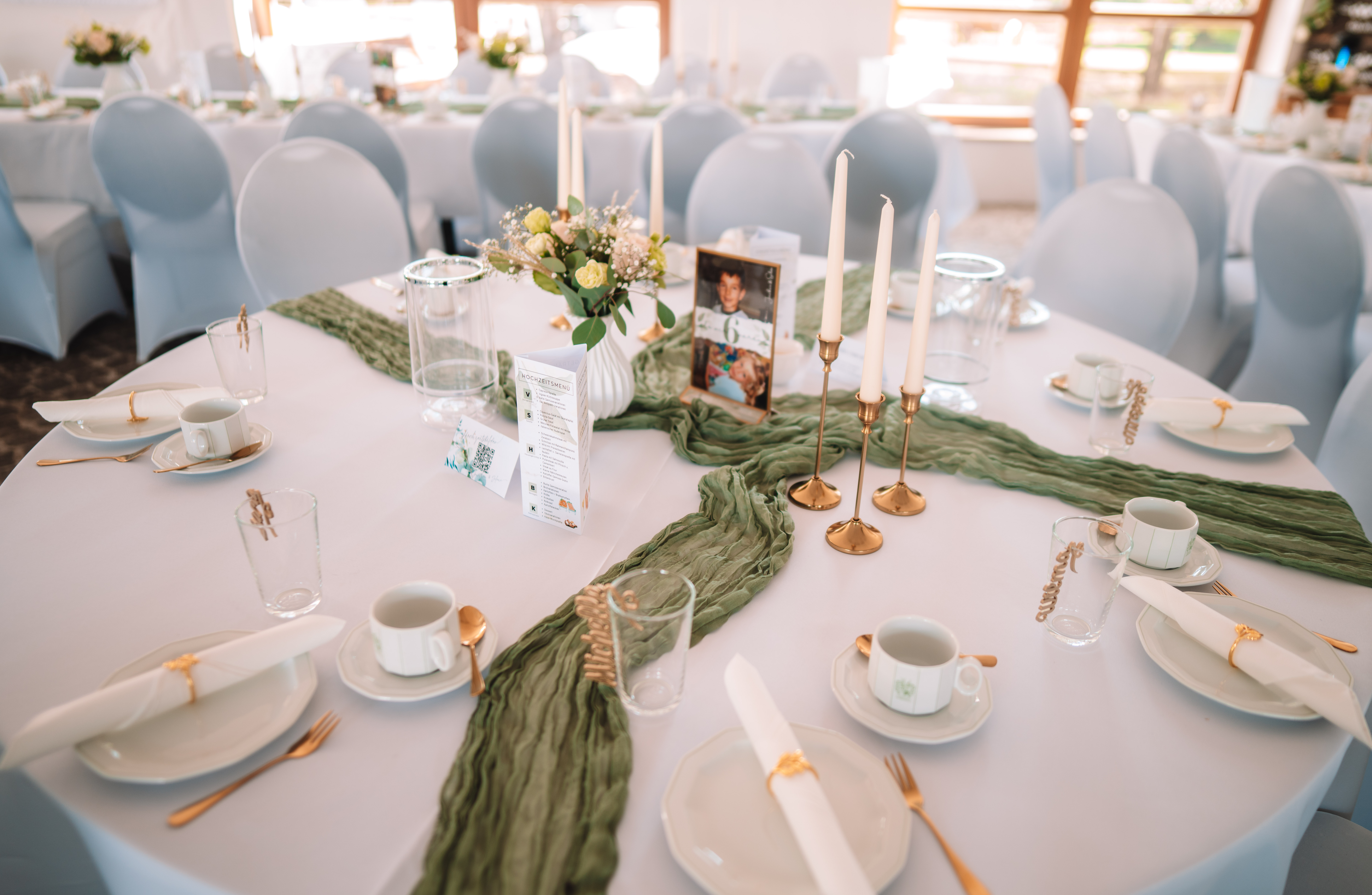 Mariage - Bennewitz - Runder Tisch mit weißer Decke, salbeigrünem Läufer, goldenen Kerzenständern, weißer Blumenvase, Menükarte und gerahmtem Kinderfoto als Tischnummer. Im Hintergrund weitere gedeckte Tische mit Hussenstühlen. - Remise am Gestüt Sachsen in Cavertitz