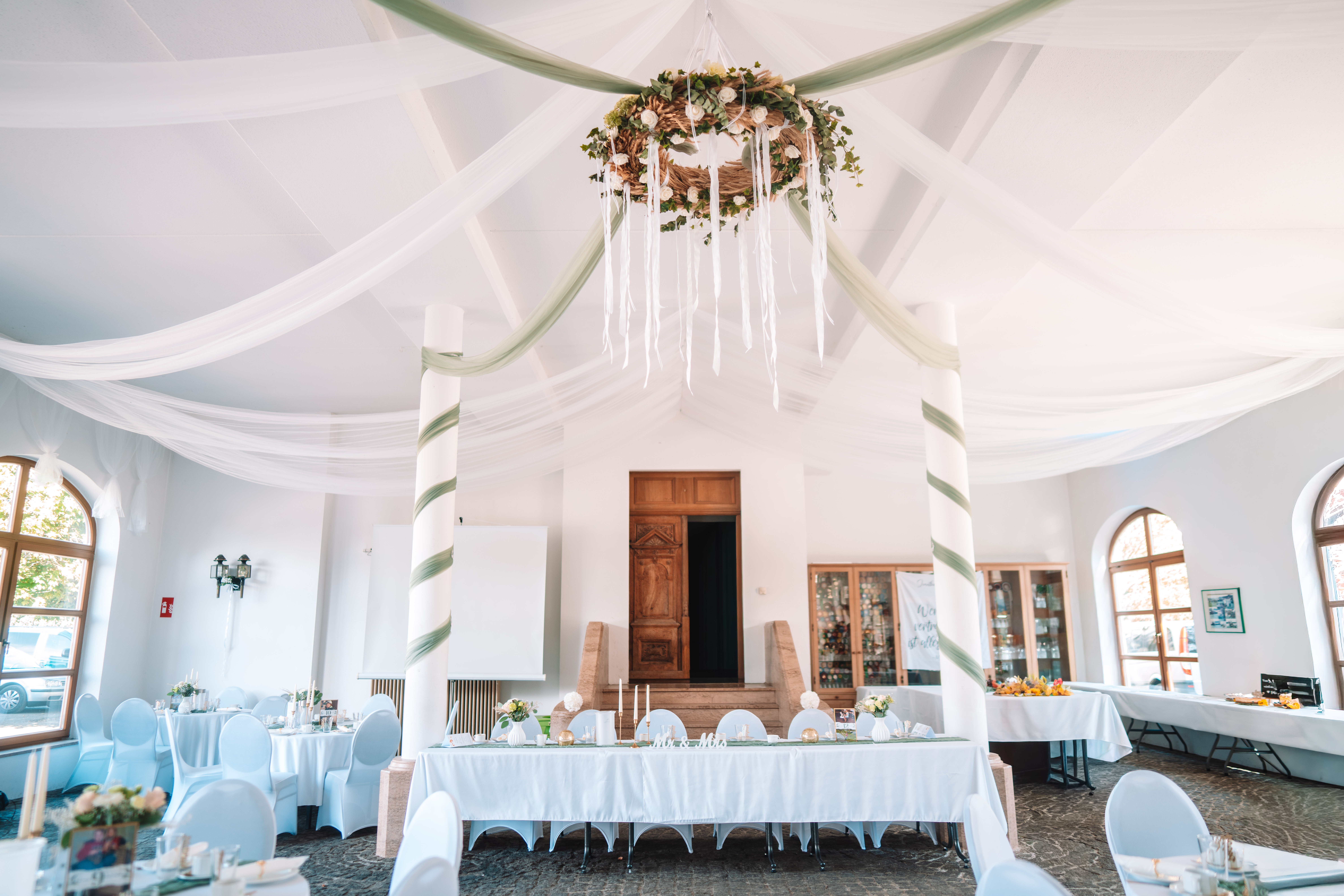 Mariage - Bennewitz - Blick unter dem dekorierten Deckenbaldachin mit Blumenkranz und Bändern hindurch auf einen alten Holzeingang, flankiert von grün-weiß umwickelten Säulen. Braut-und-Bräutigam-Tafel mit weißer Decke im Vordergrund. - Remise am Gestüt Sachsen in Cavertitz