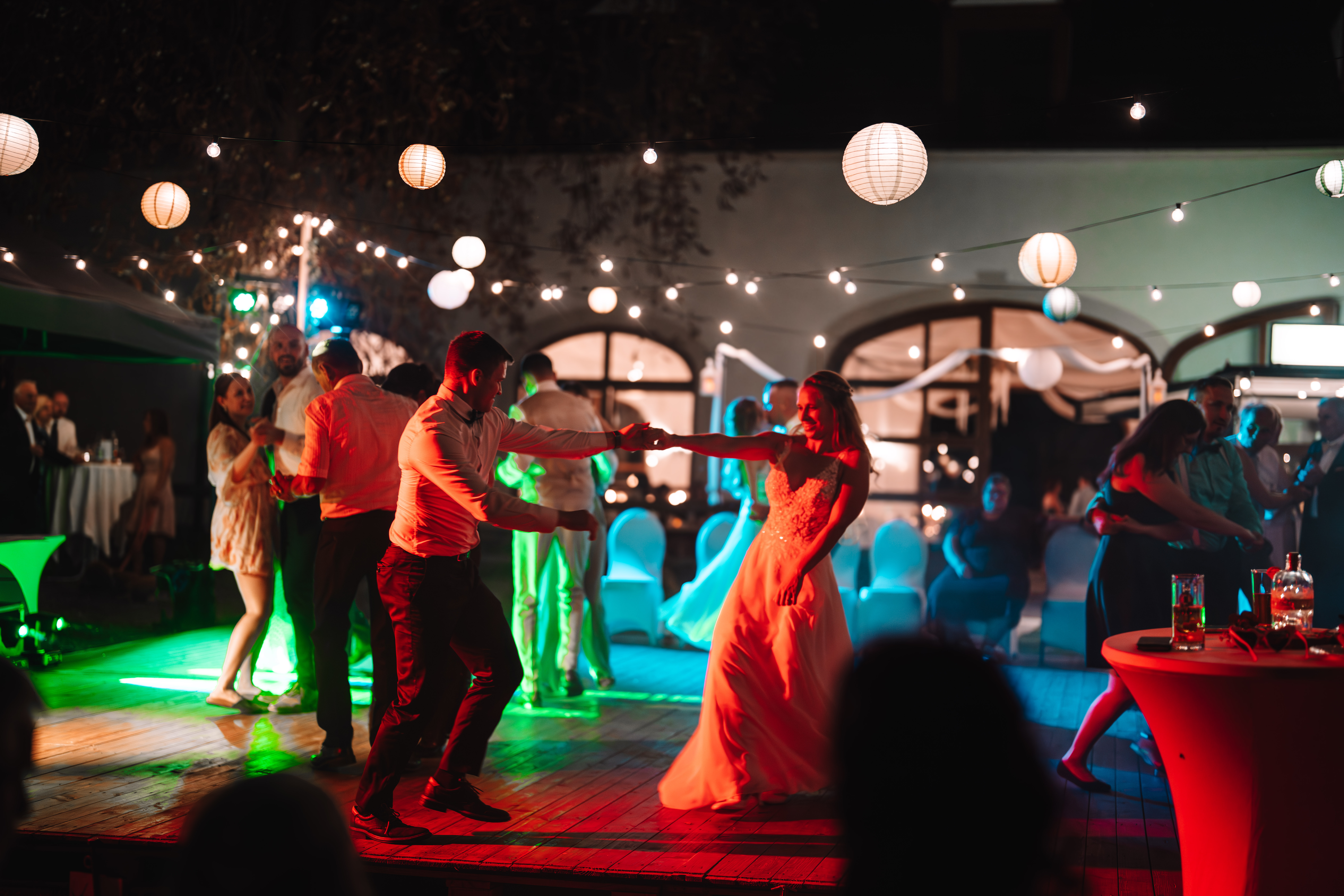 Hochzeitslocation: Nächtliche Tanzfläche mit bunten Lichteffekten (grün/rot) und Lichterketten mit Papierlaternen. Braut im orange beleuchteten Kleid tanzt mit einem Gast, weitere Paare tanzen im Hintergrund. - Remise am Gestüt Sachsen in Cavertitz