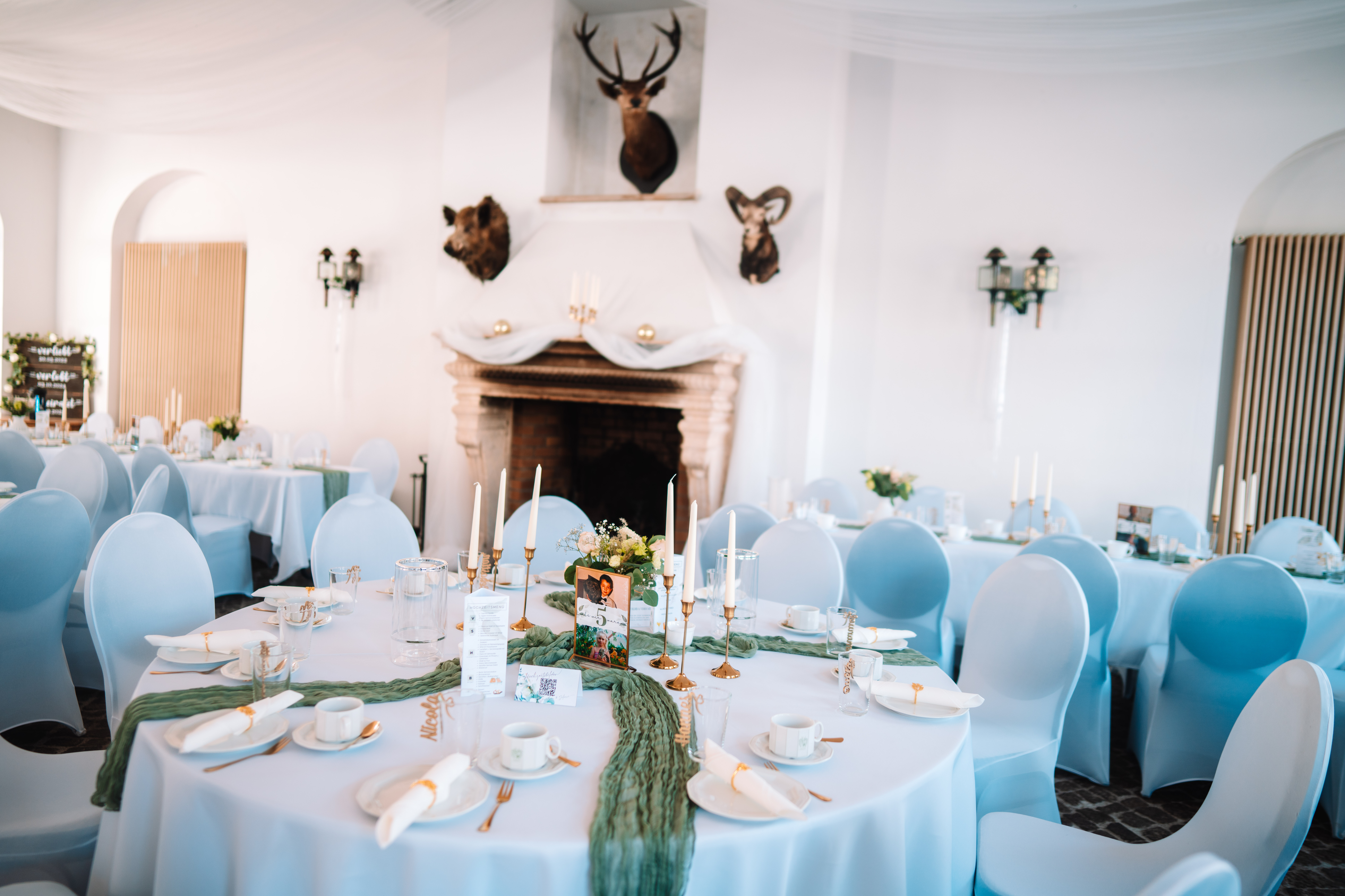 Hochzeit - Zeithain - Festsaal mit Kaminnische, darüber ausgestopfte Jagdtrophäen (Hirsch, Wildschwein, Widder). Runde Tische mit weißen Hussenstühlen, Kerzenleuchtern, grünem Läufer und Tischnummer „5" mit Foto. - Remise am Gestüt Sachsen in Cavertitz - JS Eventdesign
